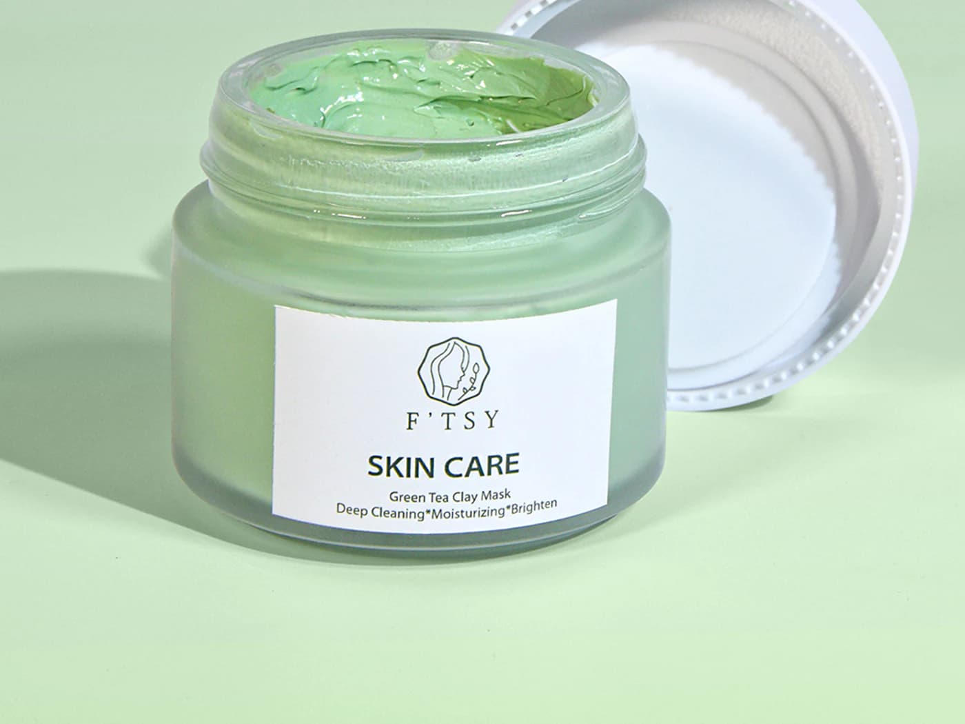 Open green clay face mask jar with the lid behind it.