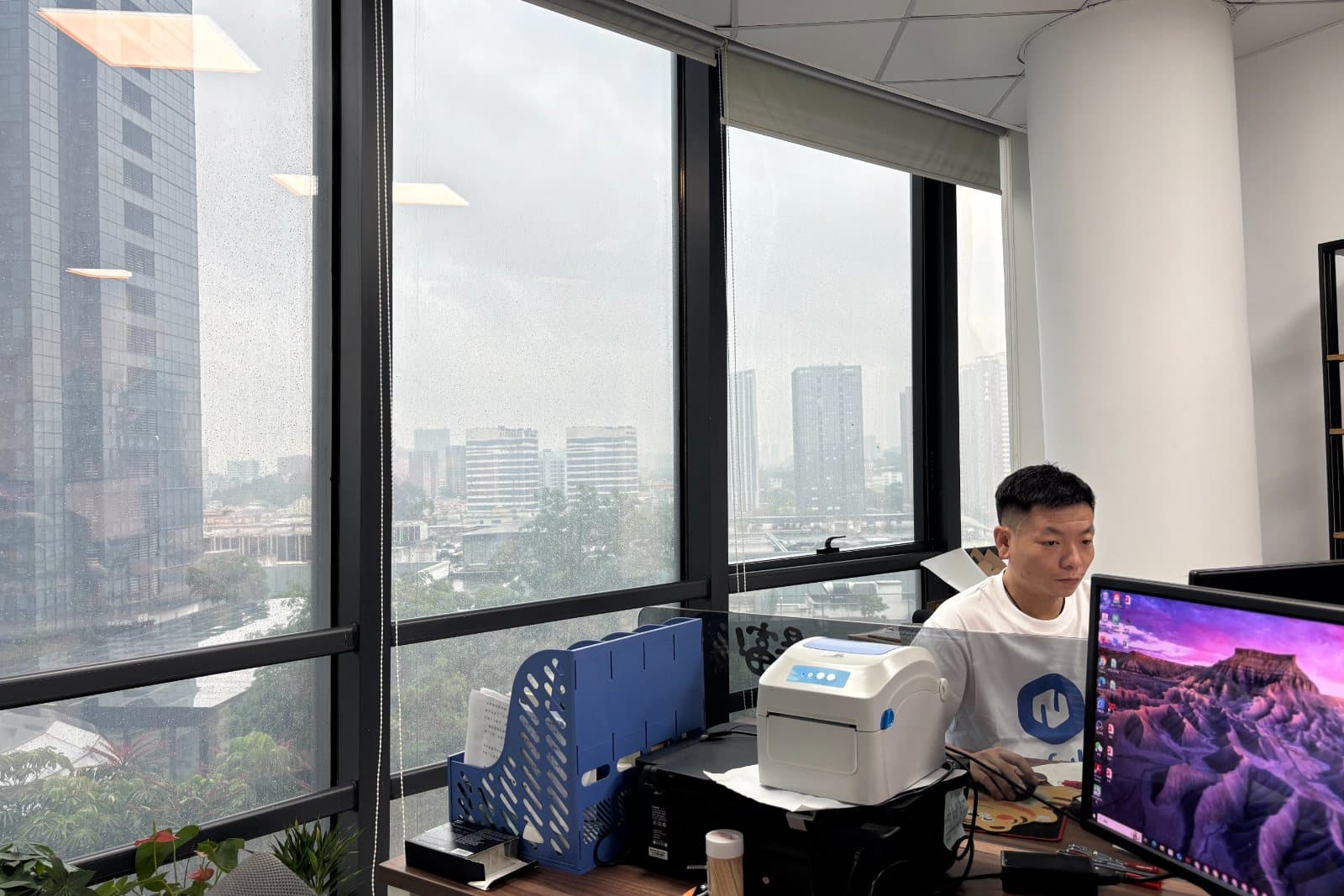 A Neofulfill team member working at a fulfillment workstation in the Shenzhen office, with a label printer and shelved products behind.